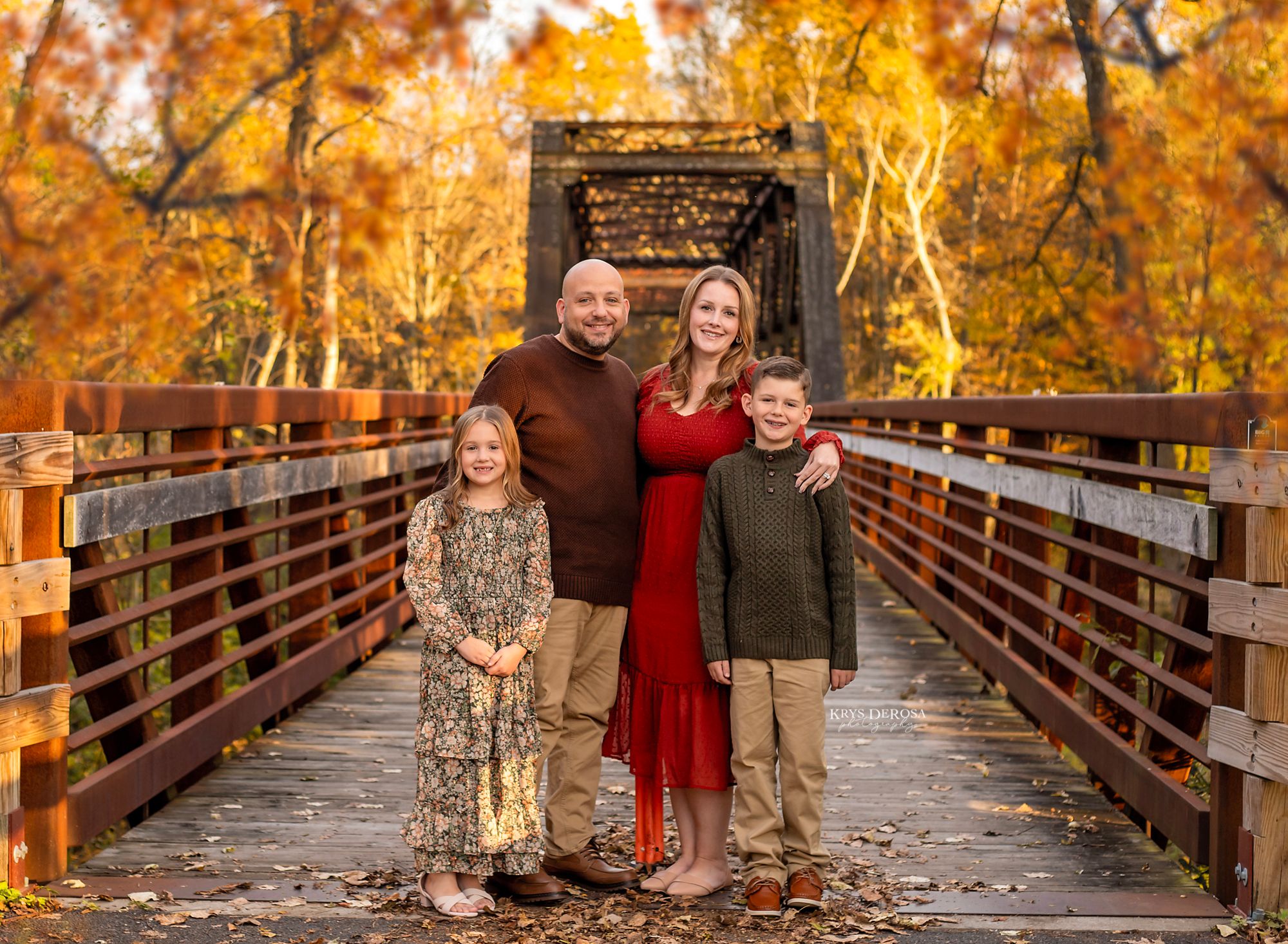 Family of four posing on a wooden bridge surrounded by vibrant autumn foliage for a fall portrait.