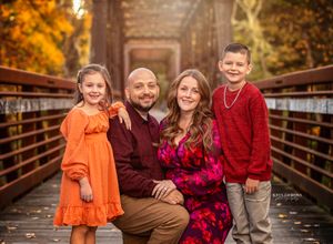 A family poses together on a wooden bridge during autumn with fall foliage in warm orange and golden colors.