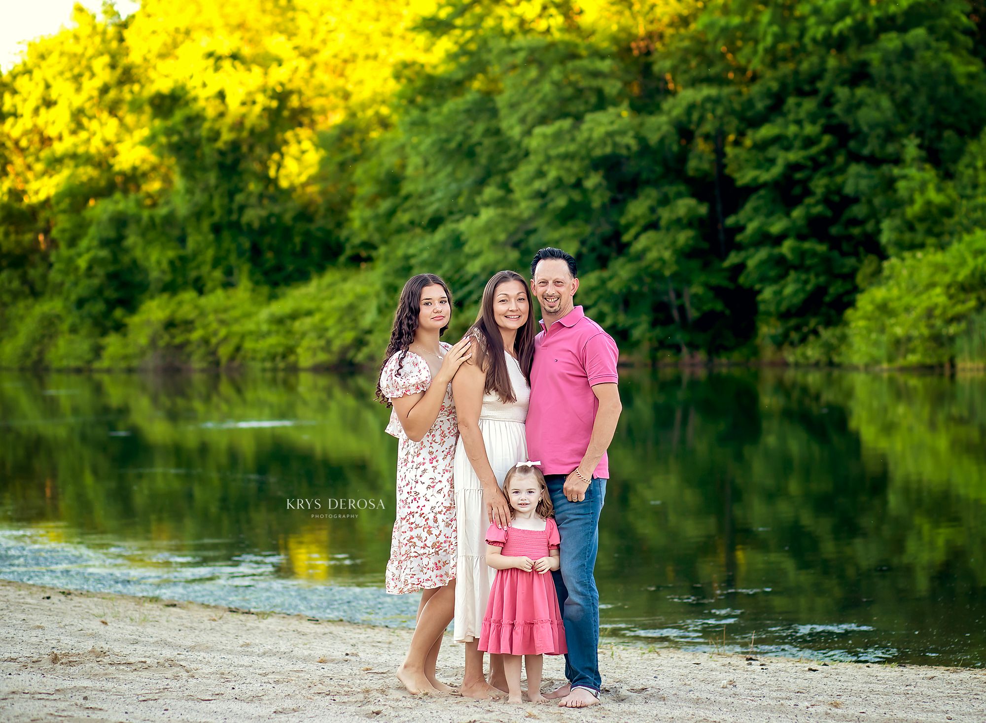 Family of four posing by a serene river surrounded by lush green trees during golden hour outdoor photography session.