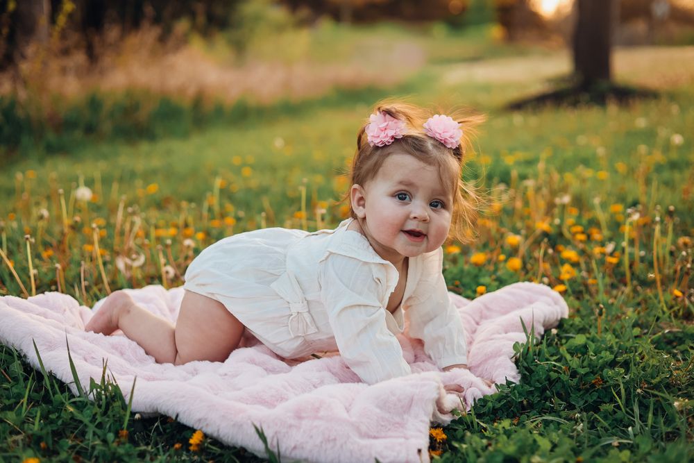 A cute baby girl in a white sweater and pink bow sits on a blanket in a sunny green meadow with wildflowers.