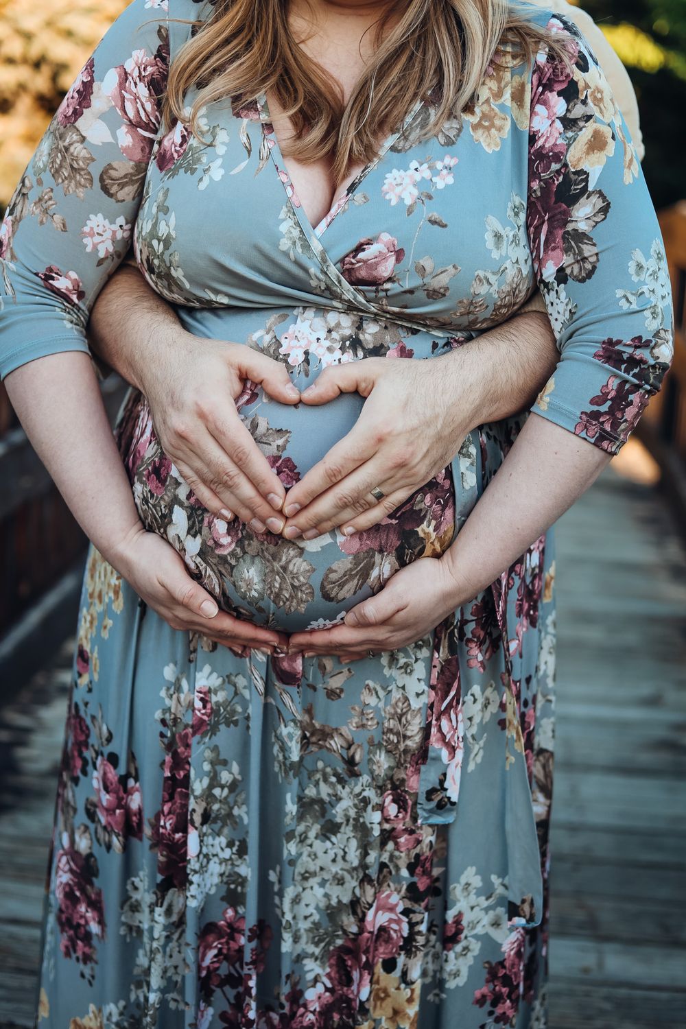 Pregnant woman in floral blue dress cradling baby bump with both hands forming a heart shape outdoors.