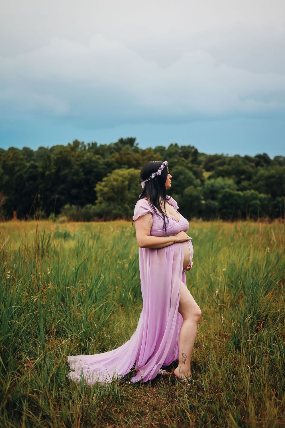 Figure in flowing pink dress sitting gracefully in tall grass field at golden hour. Maternity photography