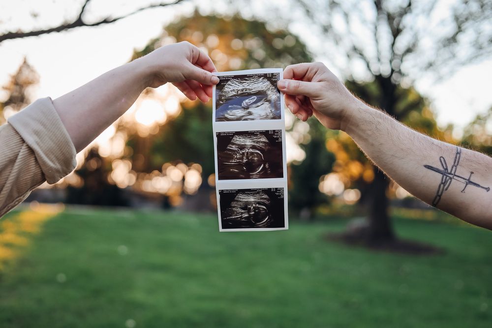 Two hands holding ultrasound photos outdoors, one with a cross tattoo, announcing pregnancy in a park setting.