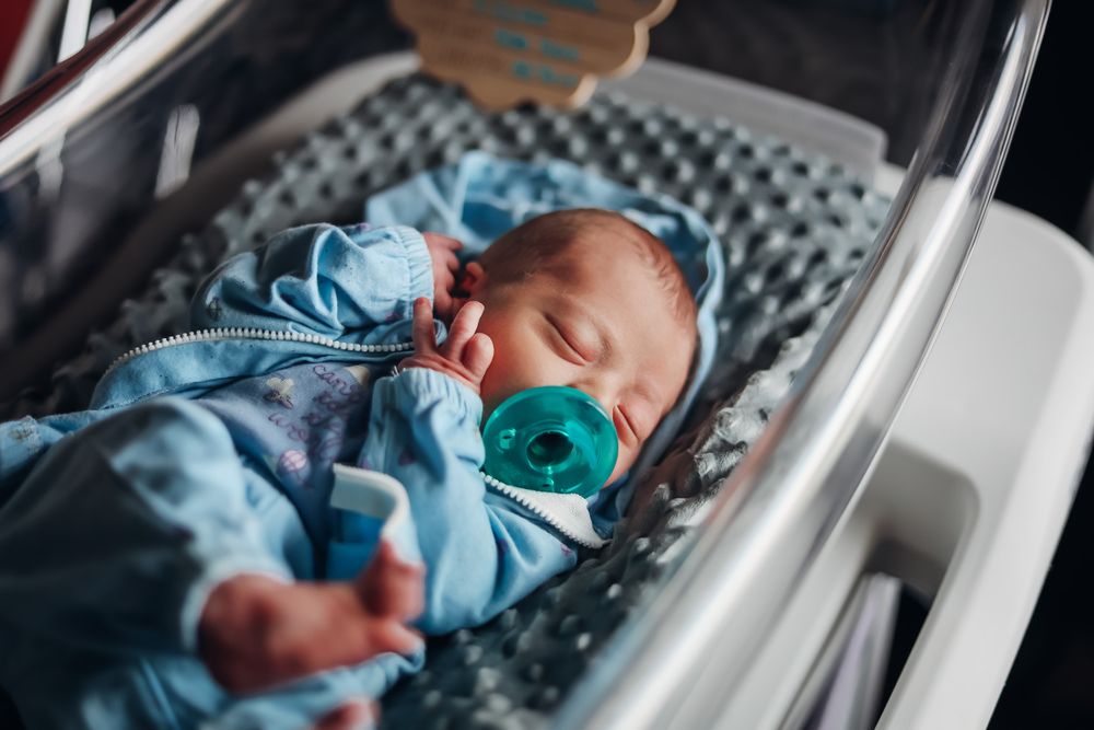 Newborn baby sleeping peacefully in hospital bassinet wearing blue onesie with teal pacifier.
