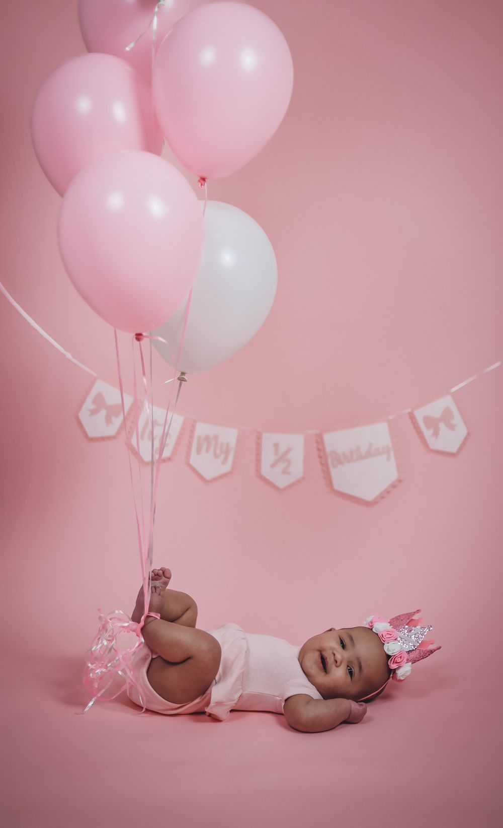 Baby in white outfit lying on pink backdrop with pink and white balloons and banner for milestone photo shoot.