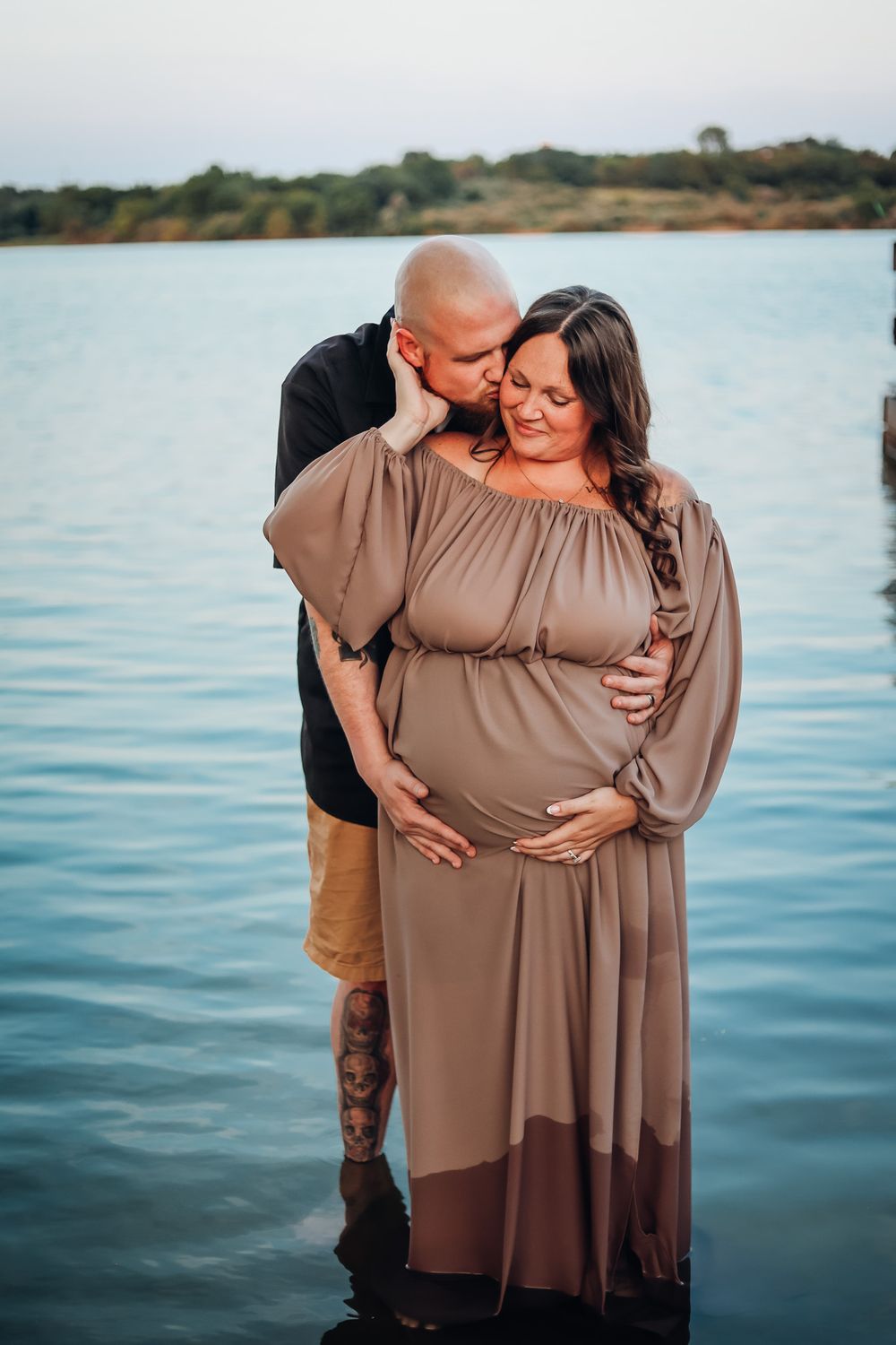 Couple shares tender moment by lake, bald man kissing pregnant woman in flowing tan maternity gown.