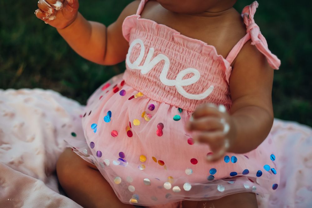 Baby girl in pink 'one' birthday dress with colorful polka dots celebrating her first birthday outdoors.