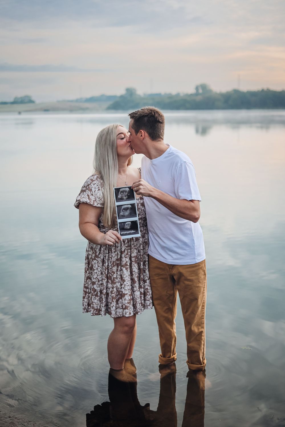 Pregnancy announcement photoshoot by calm lake at dusk with couple showing ultrasound image. Announcement photography