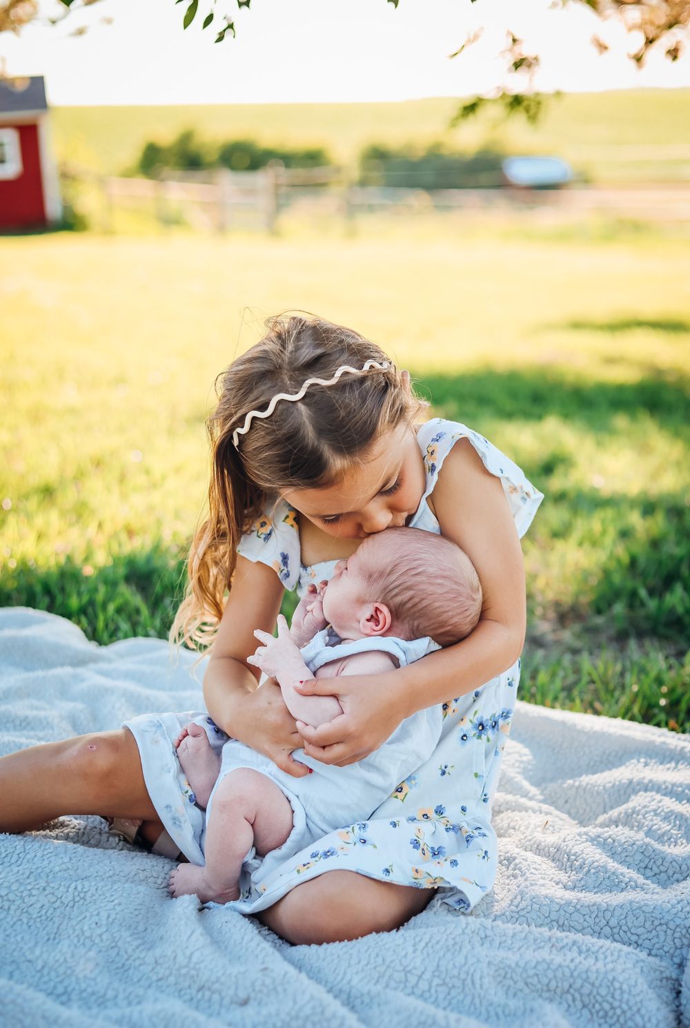 Young girl in floral dress kisses newborn baby sibling while sitting on blanket in sunny green farm field.