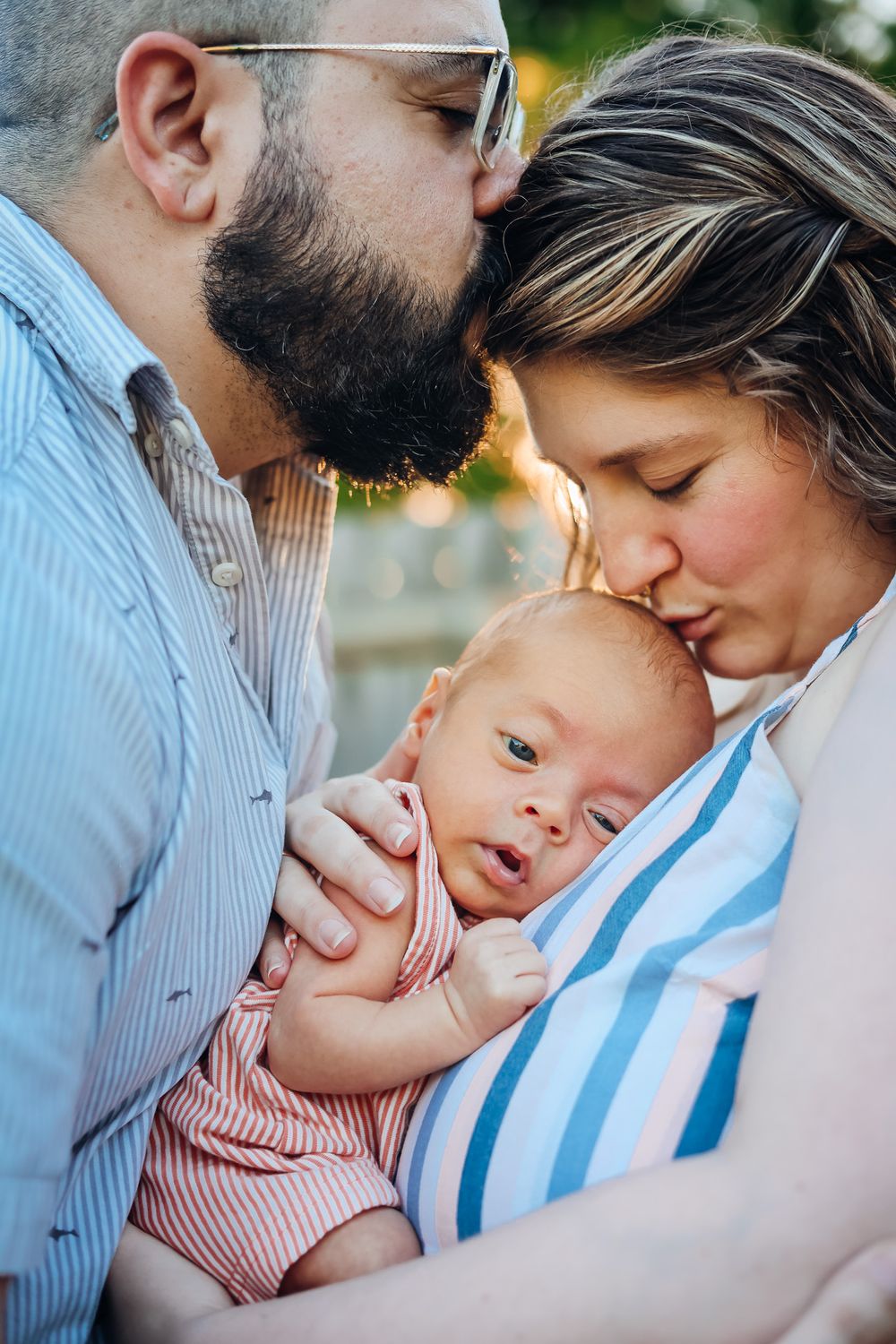 Bearded father and mother kissing their newborn baby outdoors in a tender family portrait moment.