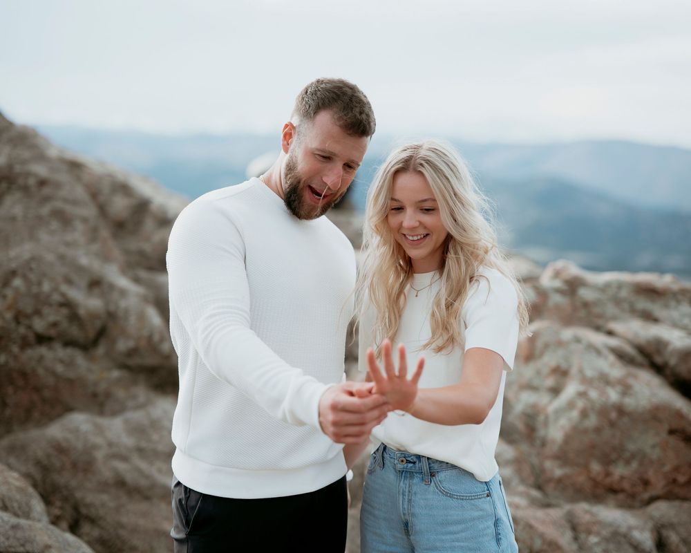 Couple in white tops examining ring together on mountain peak.
