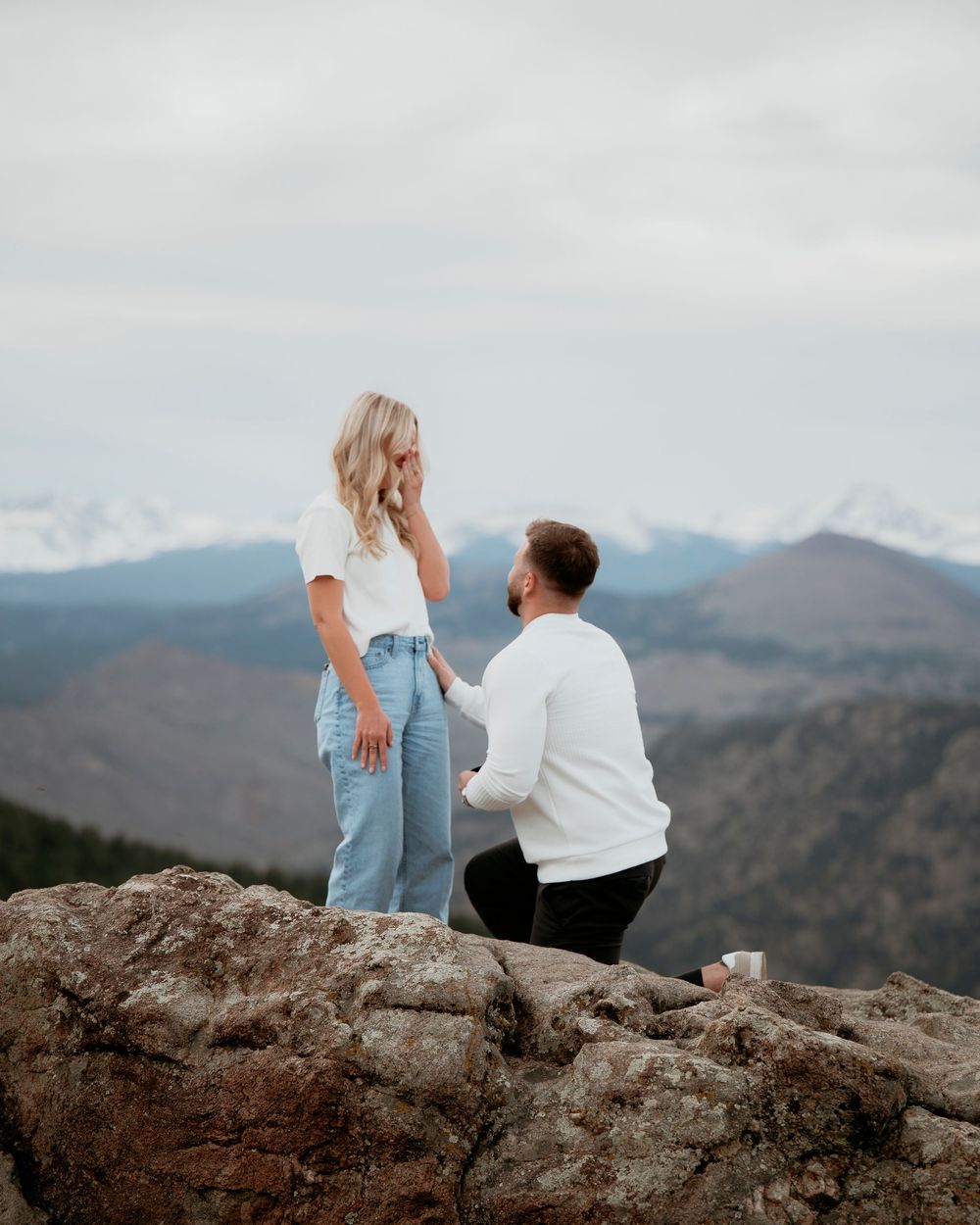 Surprise proposal moment captured on rocky mountain overlook with scenic mountain backdrop.