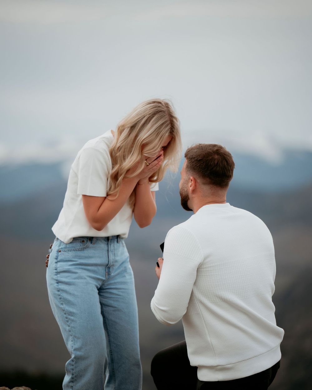 Emotional mountain top proposal with woman covering face in surprise.
