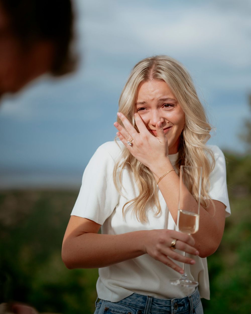 Woman with blonde wavy hair smiling emotionally while holding champagne outdoors.