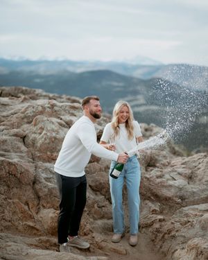 Couple celebrating with champagne spray on rocky mountain overlook during sunset.
