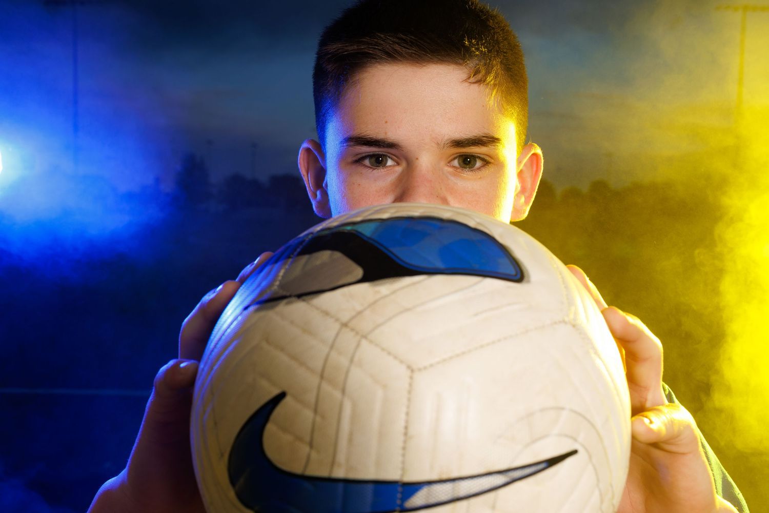 Mattoon, Illinois youth soccer player holds a soccer ball partially over his face with dramatic and smokey blue and gold lighting.