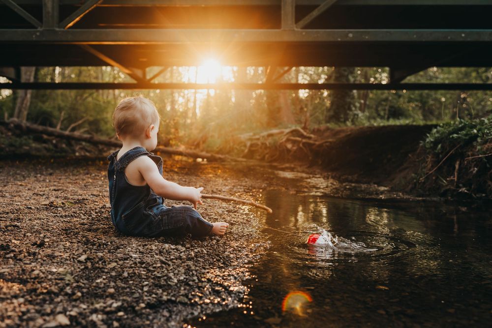 A baby sits by a sunlit creek under a bridge, playing with a toy boat in golden hour light.