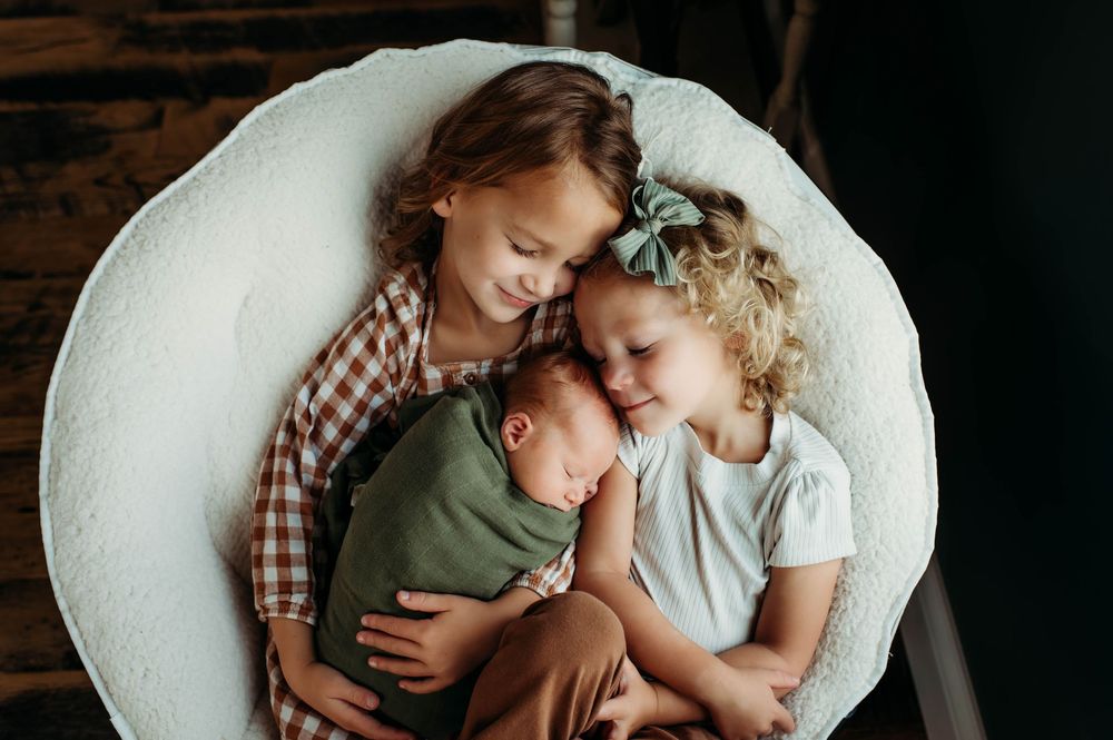 Tender family moment shows siblings and mother in muted earth tones sharing a peaceful embrace together on couch.