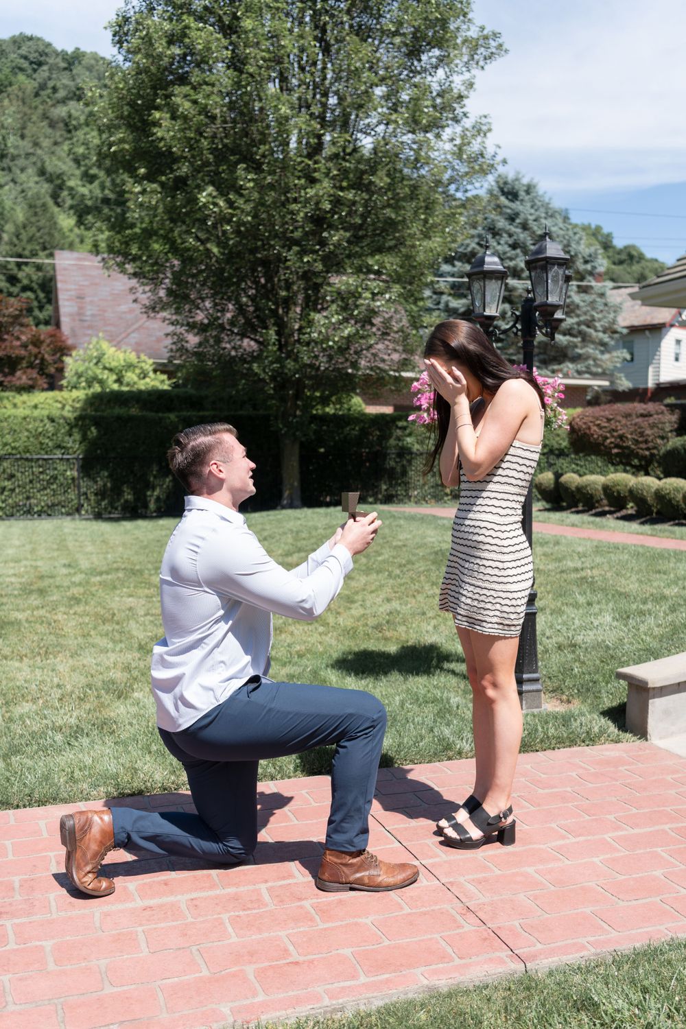 Marriage proposal moment on brick patio with man kneeling and woman covering mouth in surprise.