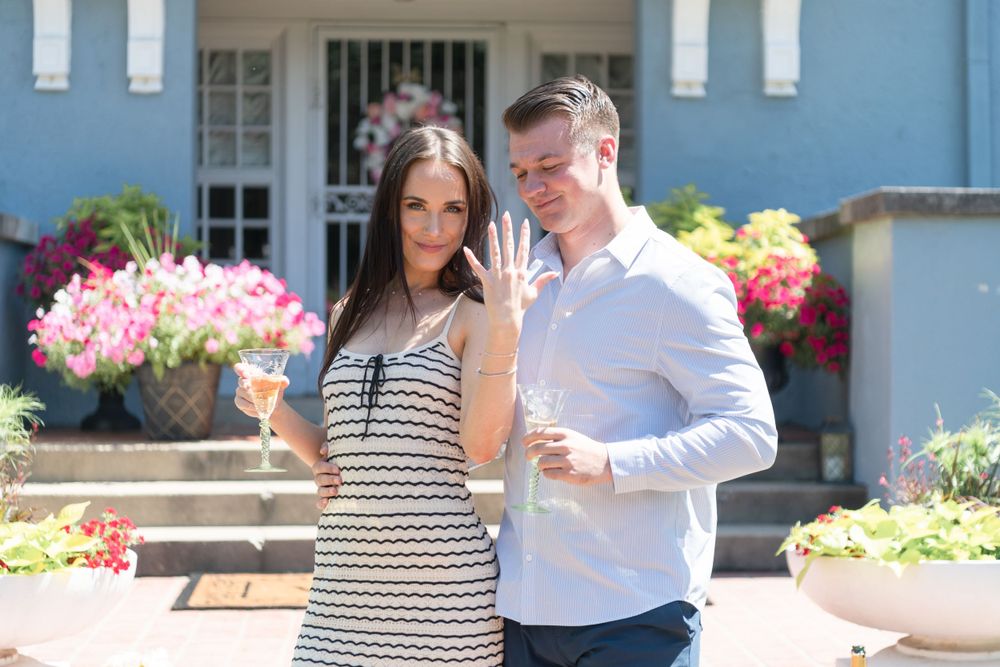 A couple celebrates with champagne on porch steps surrounded by flowers.