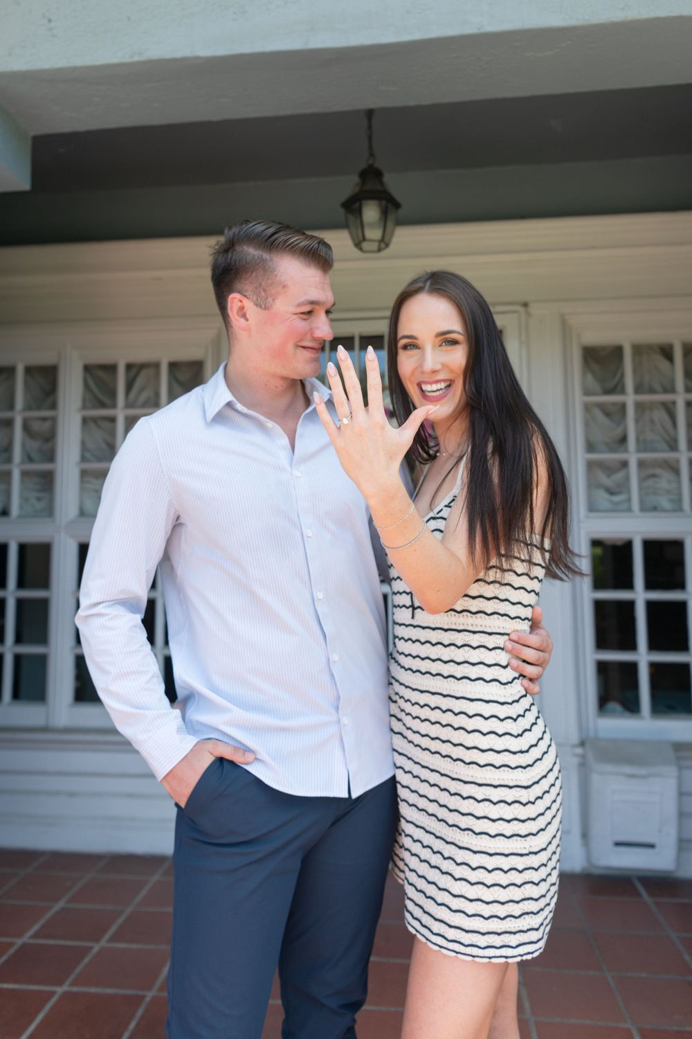 A newly engaged couple posing in front of a house entrance with traditional architecture.