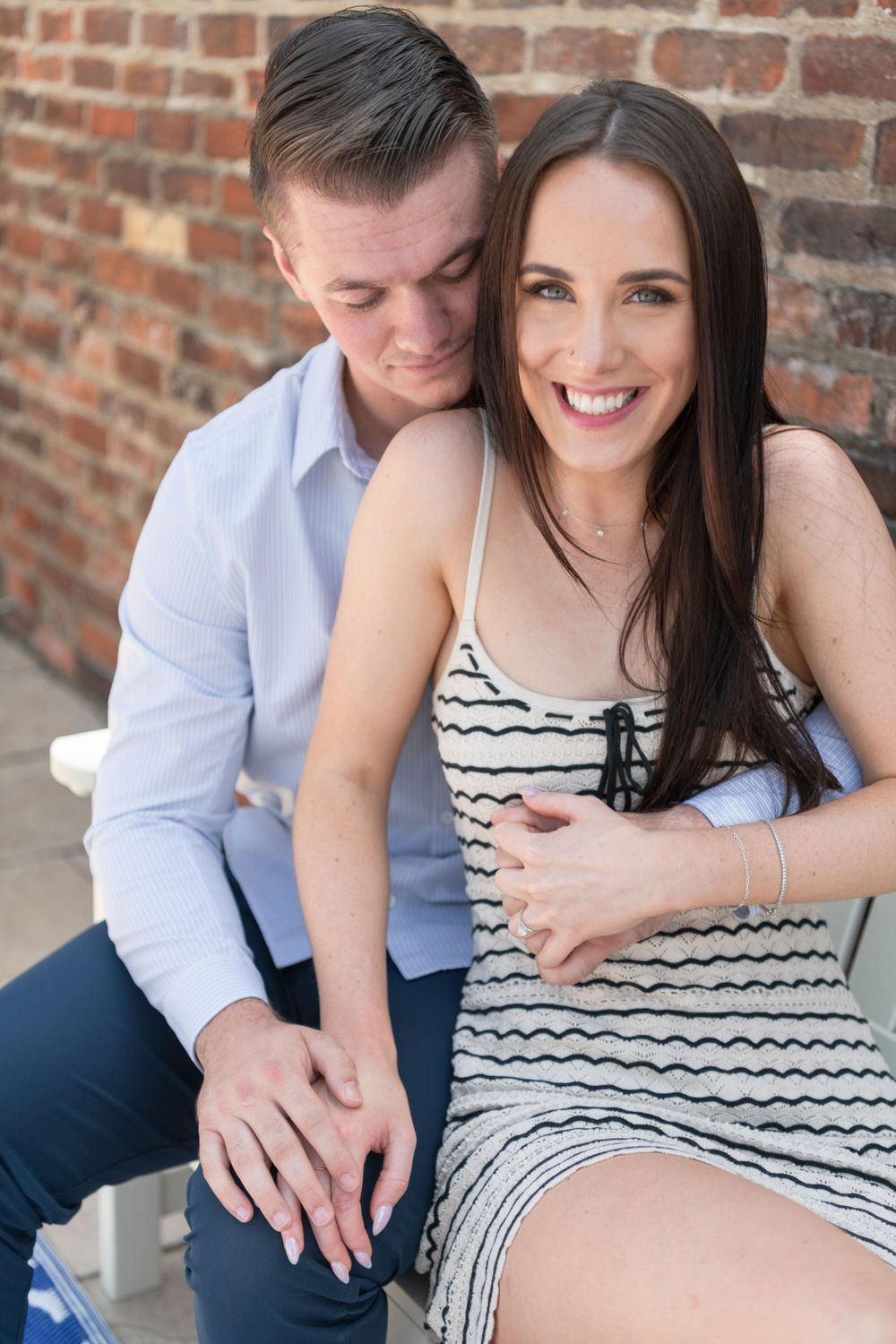 A romantic couple embraces against a brick wall, with woman wearing a striped dress and man in light blue shirt.