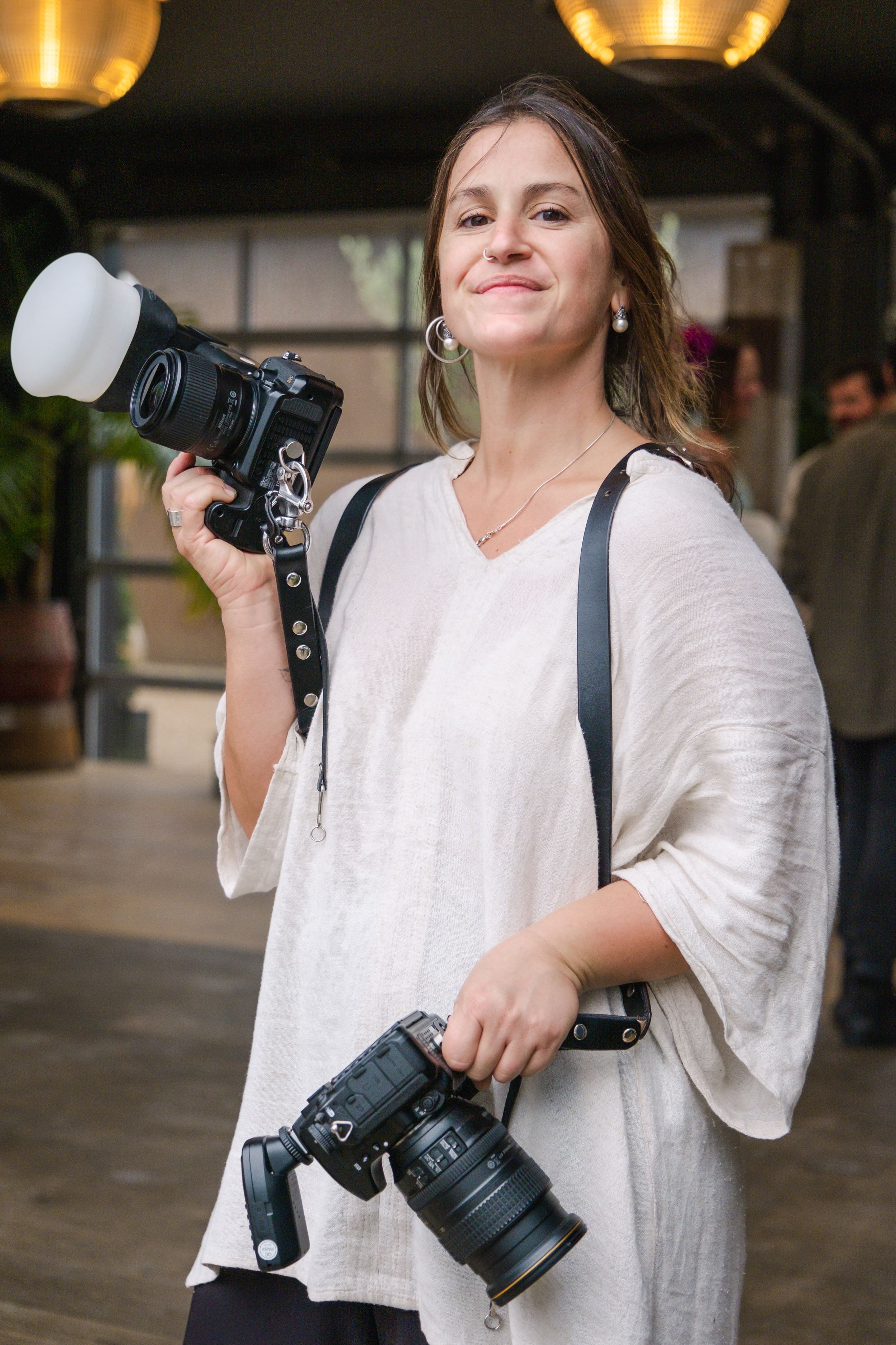 Professional photographer in a flowy white linen dress holds two DSLR cameras under warm lighting indoors.