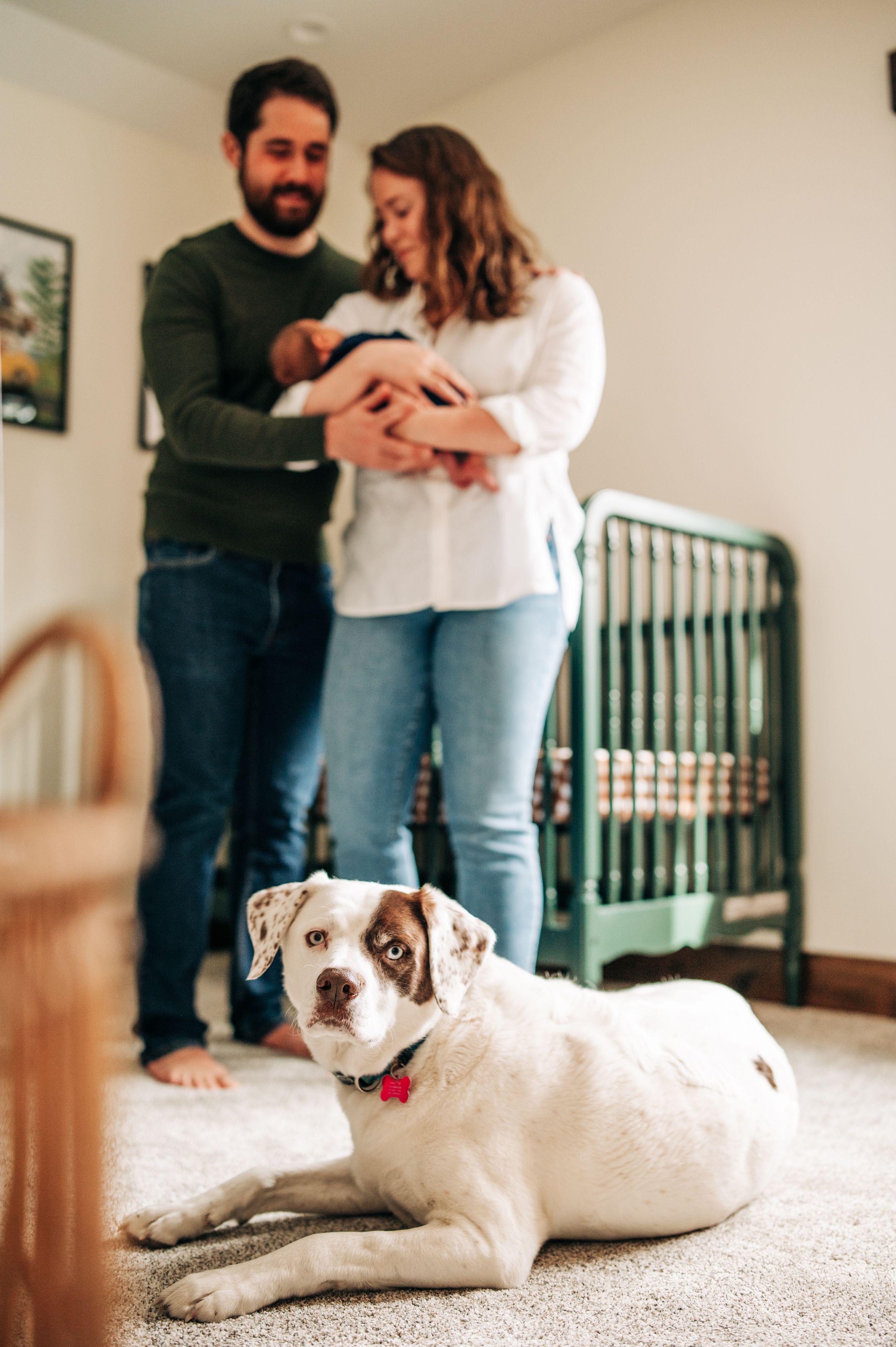 A couple stands near green crib with their white dog resting on carpet below.