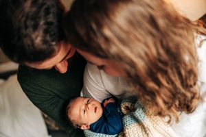 Newborn baby in blue outfit being held while parents lean in close together.