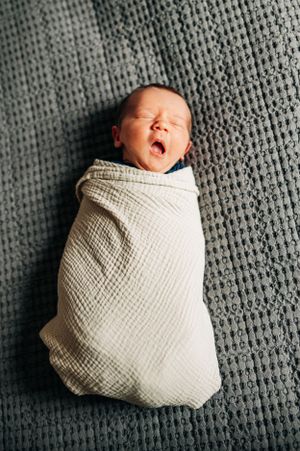 Yawning newborn baby wrapped in white swaddle on grey textured blanket.