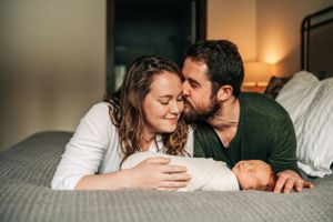 Intimate family moment with swaddled newborn on grey bed.