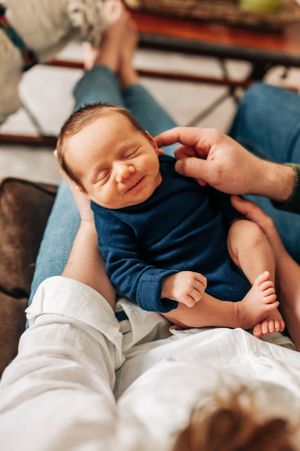 Smiling newborn baby in navy blue outfit lying on parent's lap.