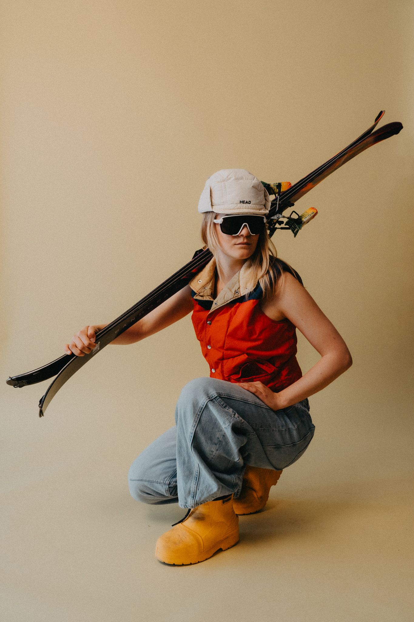 Person in orange vest and gray pants holding skis while posing against beige backdrop.