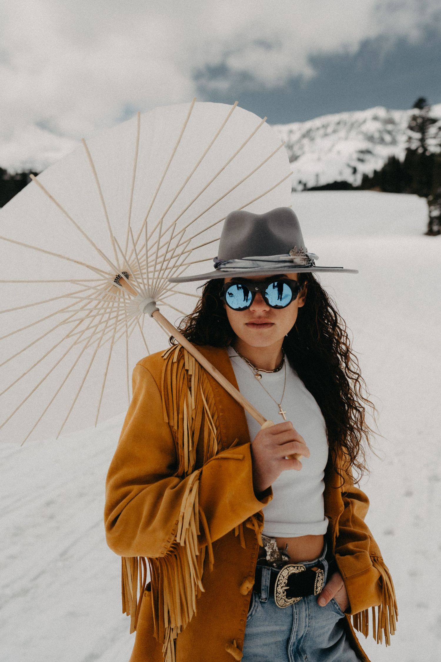 Winter fashion shoot with fringe jacket and parasol against snowy mountain backdrop.