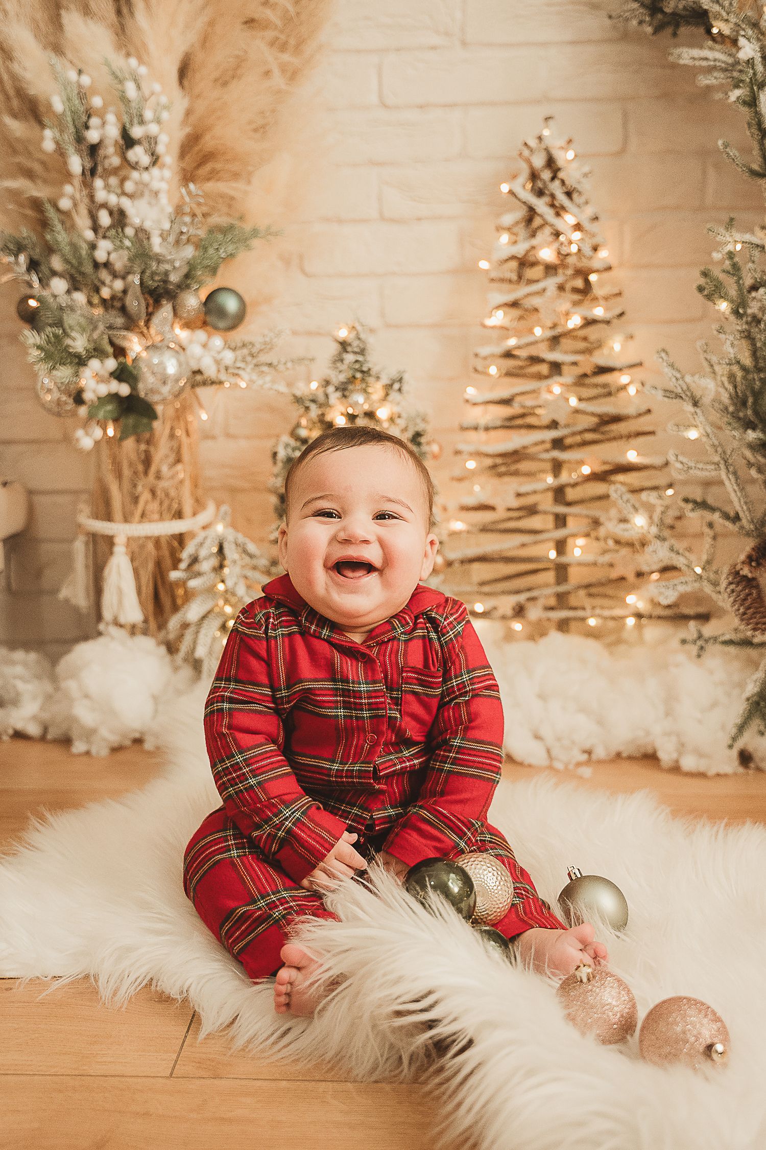 Baby in red plaid pajamas smiles during holiday photoshoot with white fur and Christmas decor.