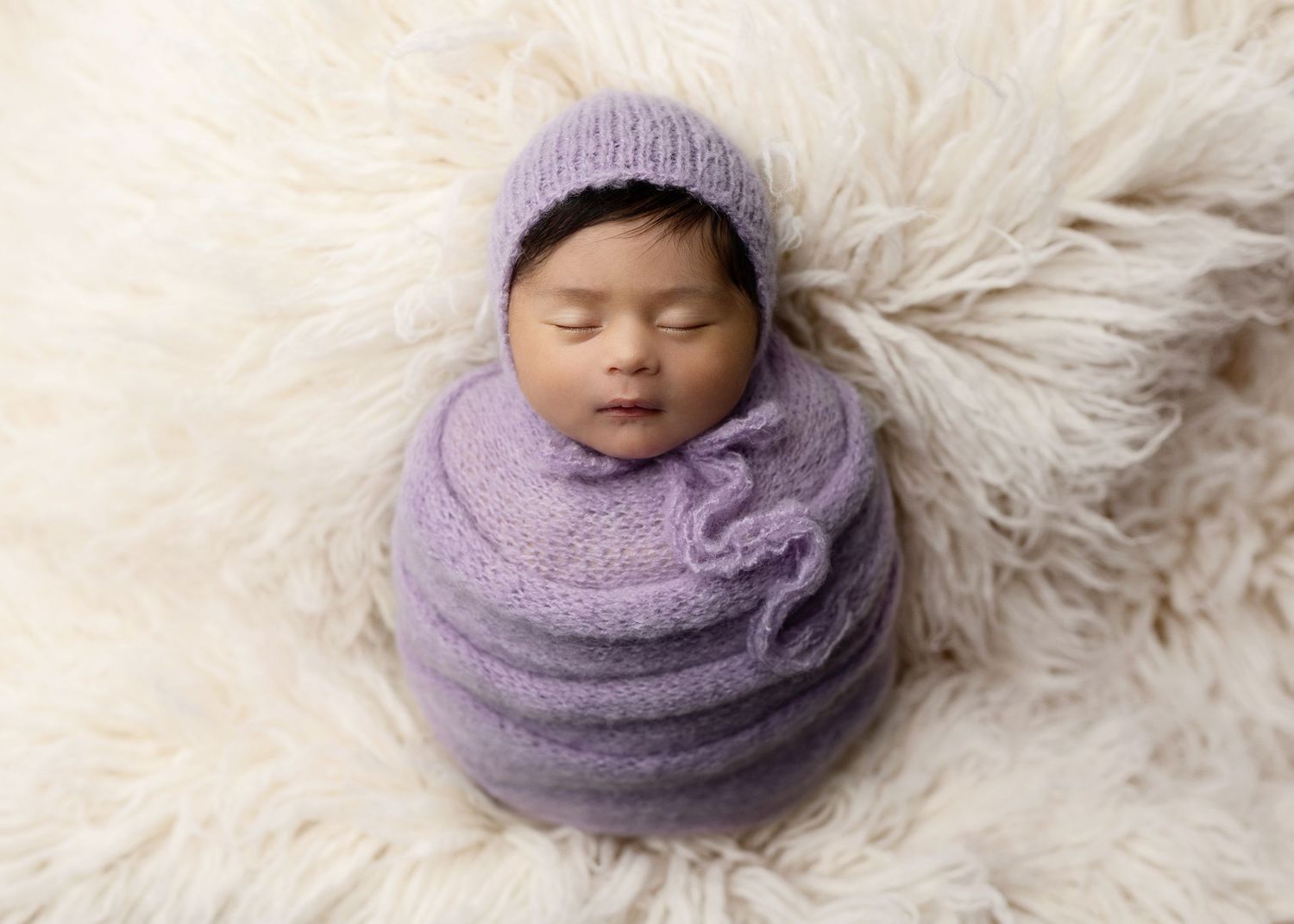 Sleeping newborn baby wrapped in purple knit fabric on a white fur background.