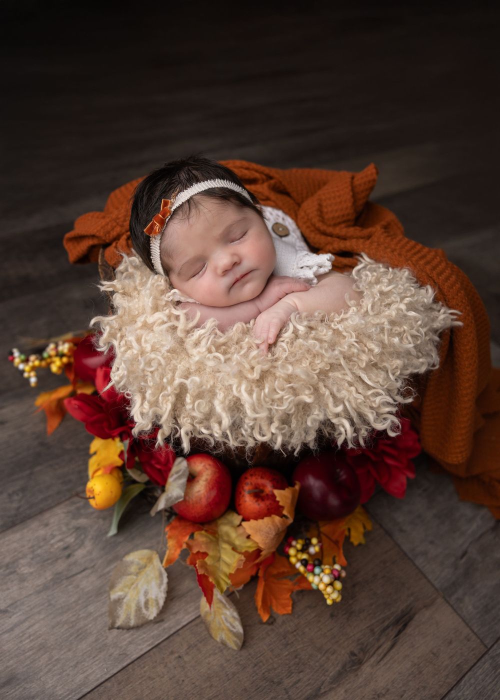 Newborn sleeps in rustic basket with autumn decorations including apples and orange blanket.
