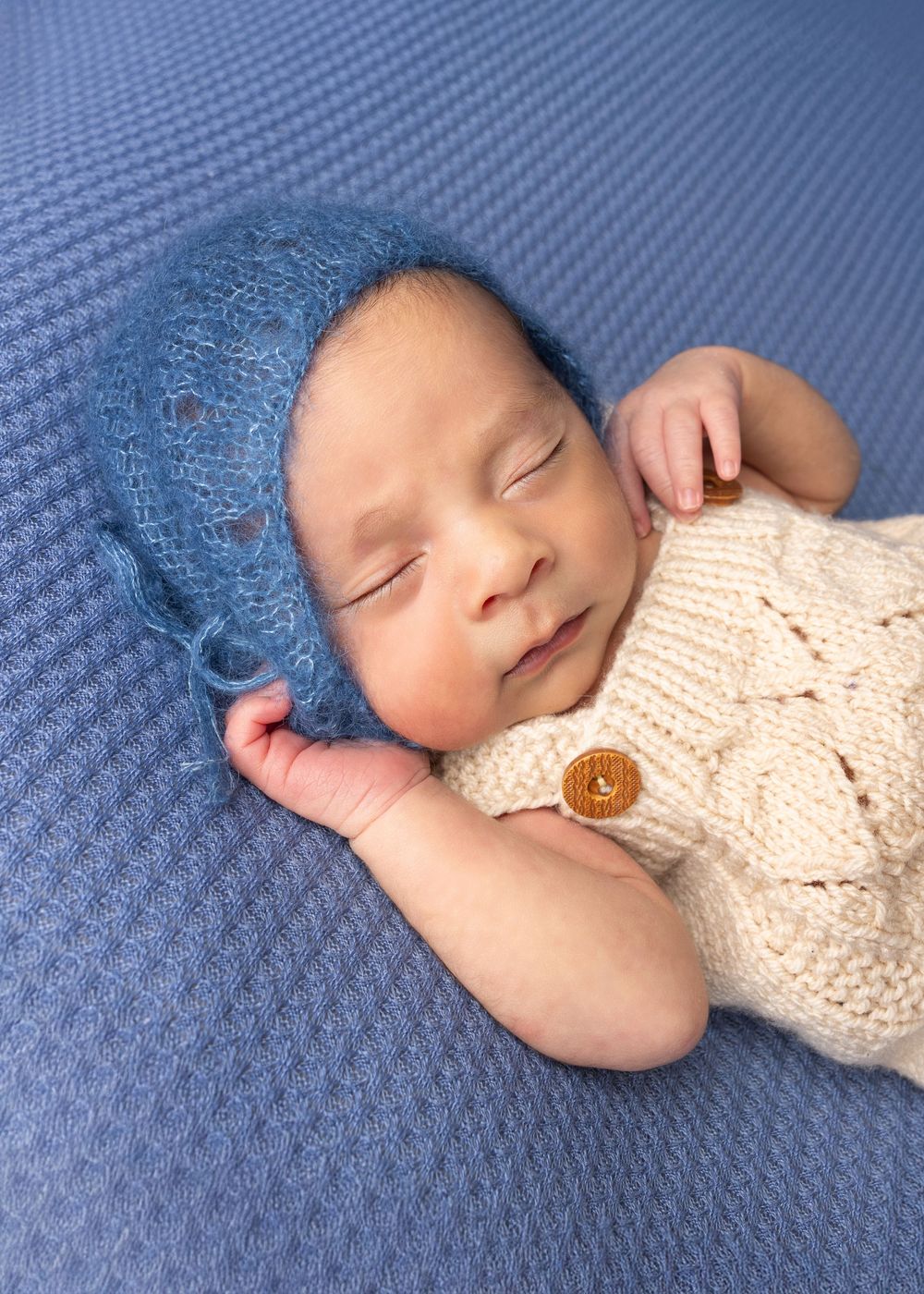 Peaceful sleeping newborn wearing blue knit bonnet and cream sweater rests on textured blue background.
