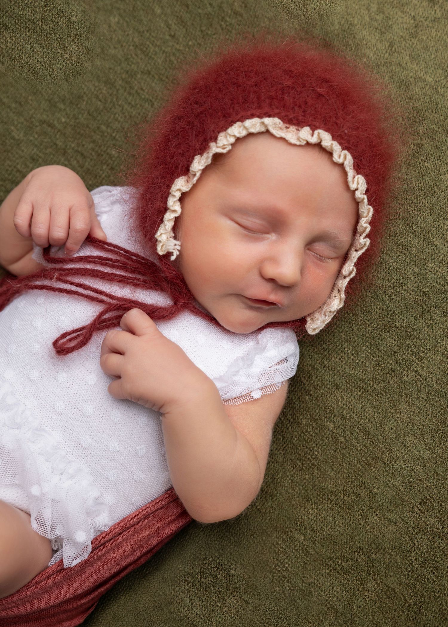 A sleeping newborn wearing a red bonnet and white outfit lies on a green textured surface.