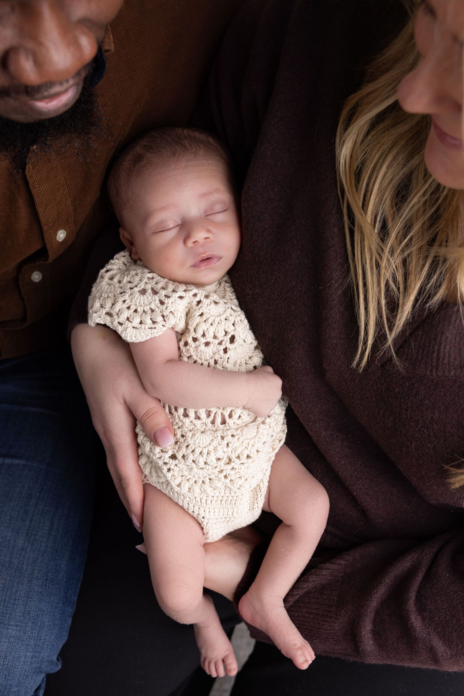 A sleeping newborn baby wearing a cream-colored crochet outfit is cradled between two people.