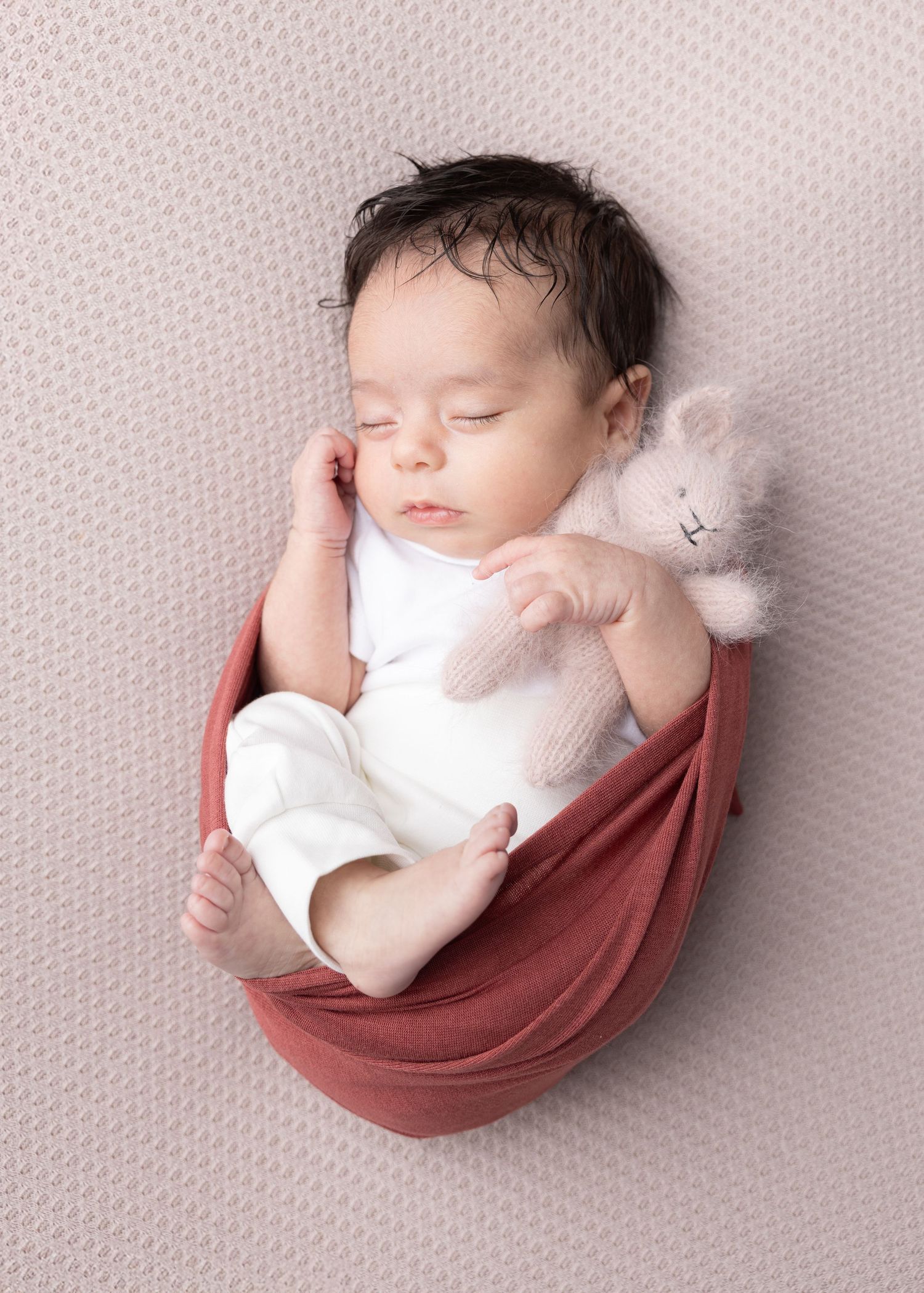 A sleeping newborn baby swaddled in white lies peacefully in a curved dusty rose fabric nest against a textured backdrop.