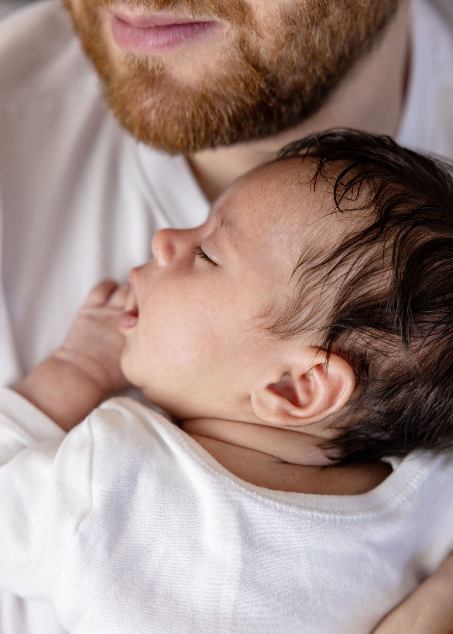 A father and baby share a tender moment while lying together on white bedding.