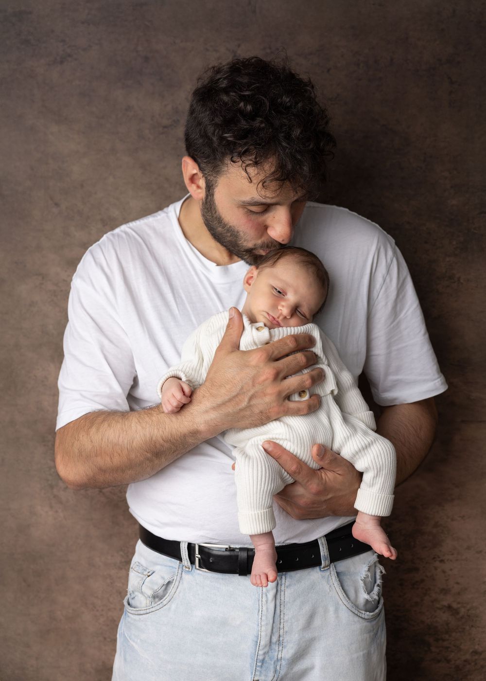 A tender moment captured as a parent in white t-shirt and jeans cradles a sleeping infant in white clothing.