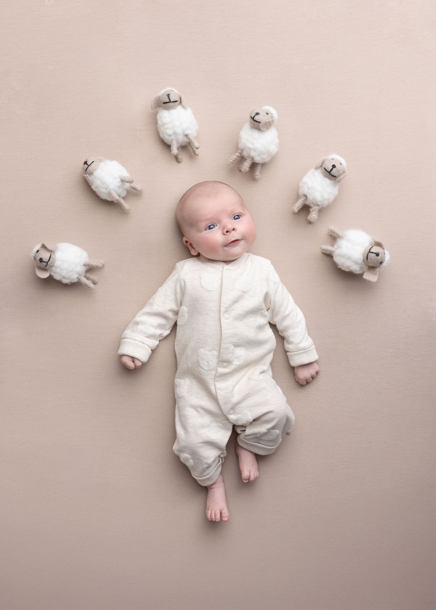 A baby in white sleepwear lies on beige background surrounded by scattered white toilet paper rolls.
