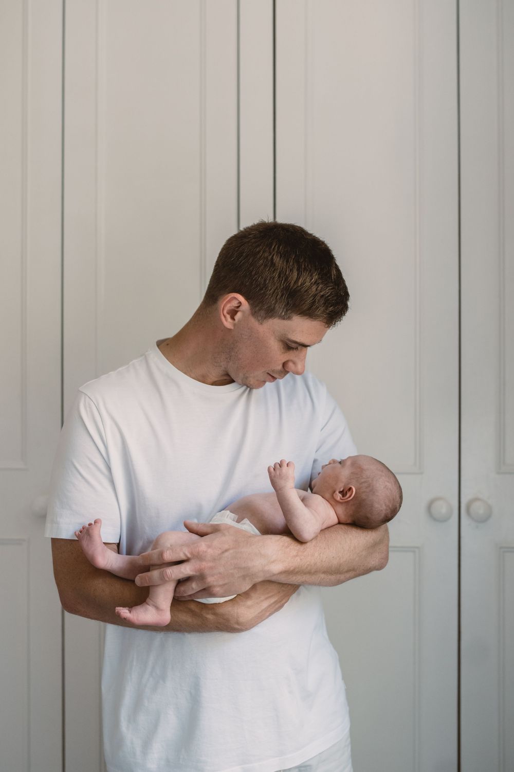 Father in white shirt cradles and bonds with newborn baby against light gray wall background.