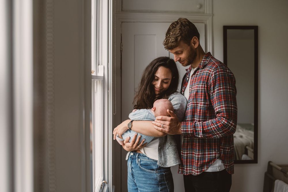 A couple stands by a window in natural light while tenderly holding their newborn baby.