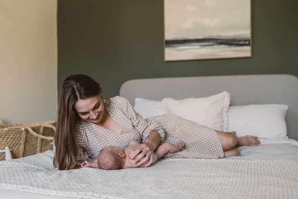 An intimate lifestyle photograph showing a tender moment between mother and newborn baby on a white bed.