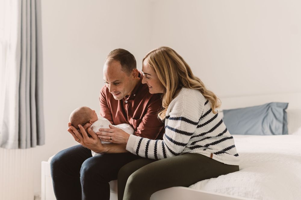 Parents sitting together on a bed, tenderly holding a newborn baby in a bright room.