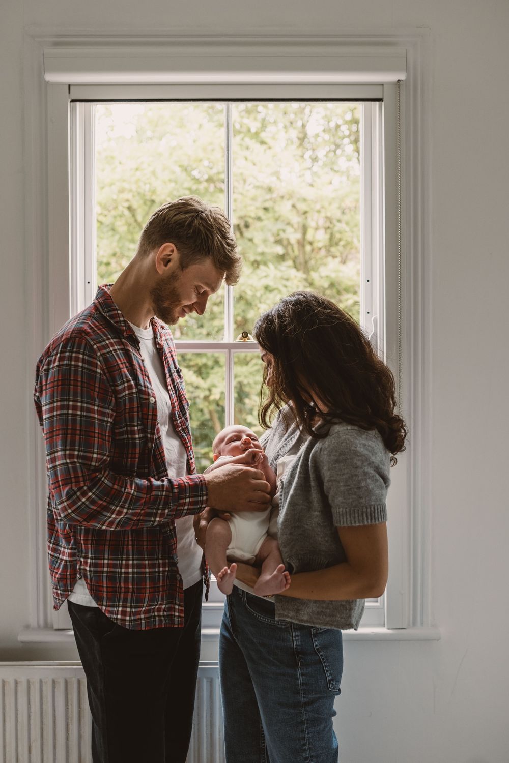 A couple in a bright room by a window shares a tender moment cradling their newborn baby.