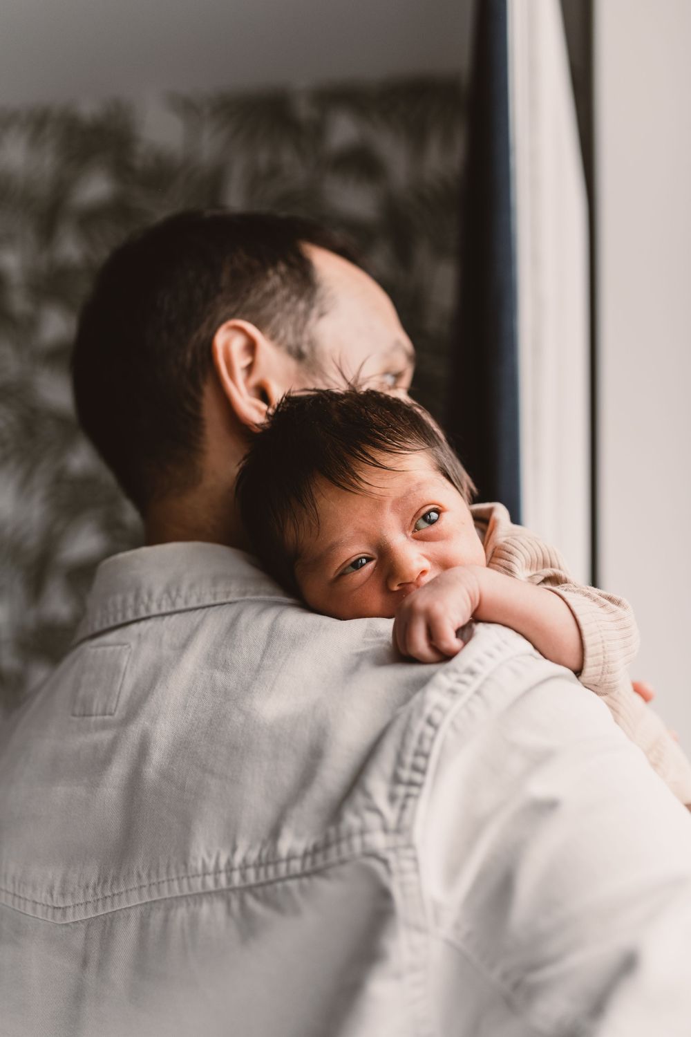 A tender moment captured as a newborn baby rests their head on his father's shoulder in warm, natural lighting.